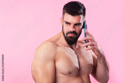 Shirtless strong bodybuilder talking on his cell, using a smartphone, pink studio background
