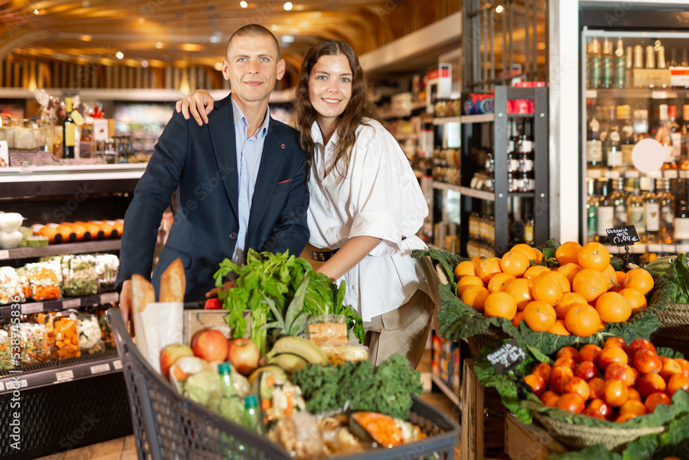 Portrait of young woman and man with shopping cart at vegetables department at supermarket