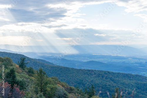 Rays of light can be seen shining through the clouds over the blue Ridge Mountains inside Shenandoah National Park. The foreground of the photo features the brush and forests of a mountain.