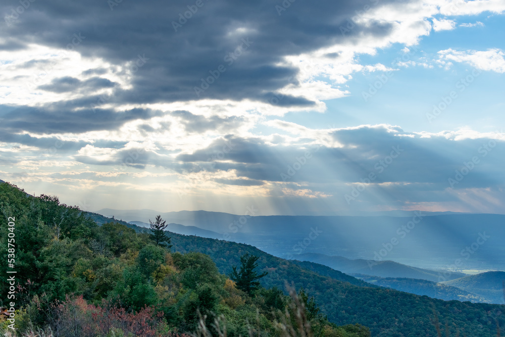 Rays of light can be seen shining through the clouds over the blue Ridge Mountains inside Shenandoah National Park. The foreground of the photo features the brush and forests of a mountain.