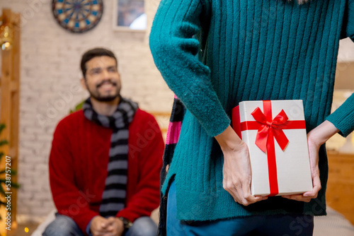 Rear view young caucasian woman hide presents for surprise boyfriend in celebration Christmas at home, woman holding gift hide behind for giving man with excited, xmas and new year or holiday concept.