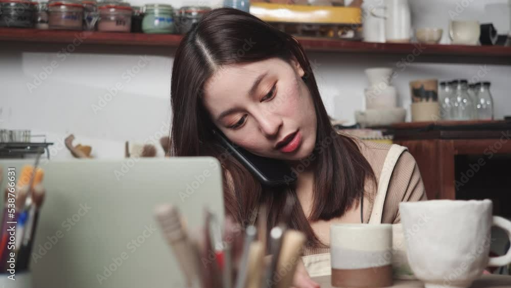 Young woman using smartphone and receive orders clay ceramic from customer. Woman typing keyboard on laptop computer for real time sell in business. Concept of business, technology and logistic. Young woman using smartphone and receive orders clay ceramic from customer. Woman typing keyboard on laptop computer for real time sell in business. Concept of business, technology and logistic.