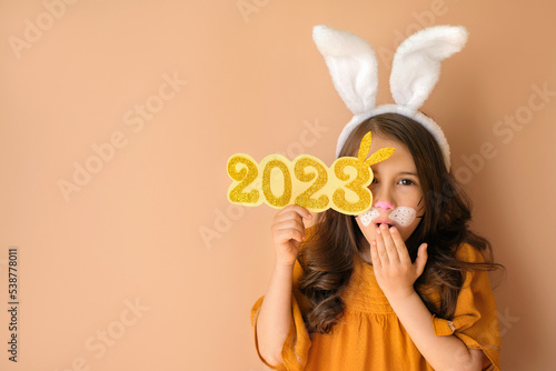 An attractive gorgeous girl in guise of rabbit holds number 2023, covering her mouth open in surprise with hand. New Year's discounts, sales and promotions.