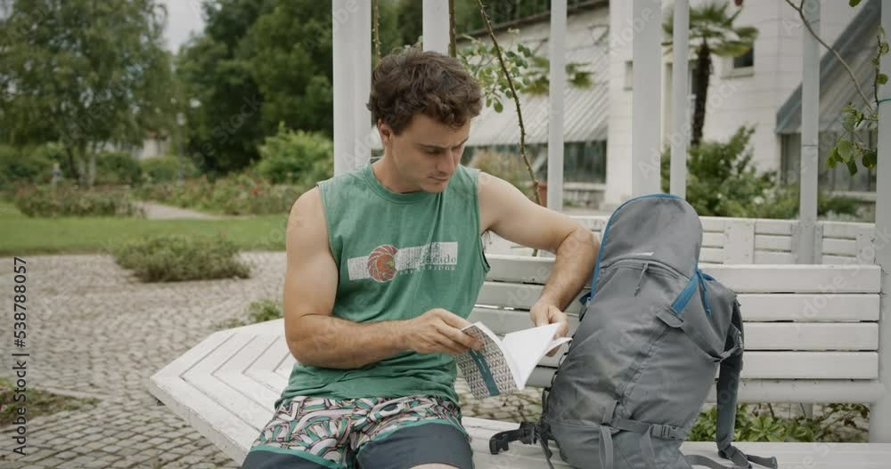 Young man reading a booklet, sitting on a banch next to his grey backpack. Shoulders and knees exposed. Pan camera roll. Young man reading a booklet, sitting on a banch next to his grey backpack. Shoulders and knees exposed. Pan camera roll.