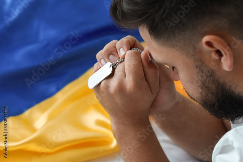 Young man with flag of Ukraine and military tag praying in bedroom, closeup