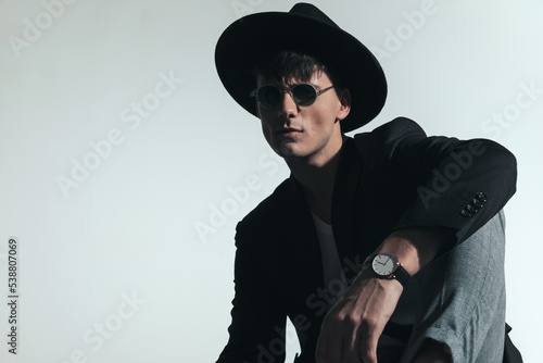 cool fashion guy with black hat holding elbow on knee and posing