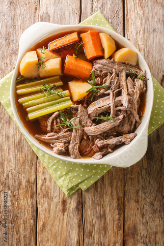delicious beef pot roast with potatoes, celery and carrots with thyme and rosemary closeup in the bowl on the wooden table. Vertical top view from above photo