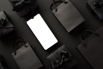Black friday shopping concept. Top view photo of smartphone black gift boxes with ribbon bows and trendy paper bags on isolated black background with empty space
