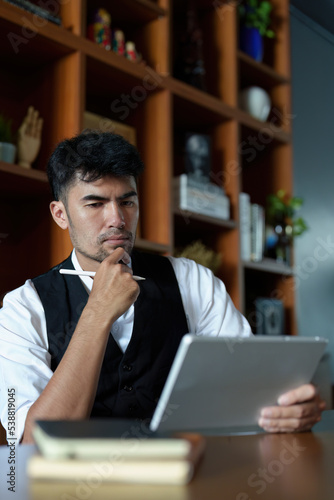 A portrait of a good looking and discreet Asian man sitting at his desk with a thoughtful look on his tablet computer