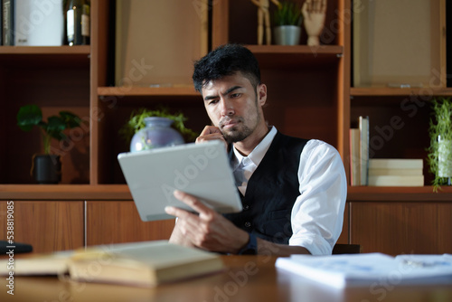 A portrait of a good looking and discreet Asian man sitting at his desk with a thoughtful look on his tablet computer