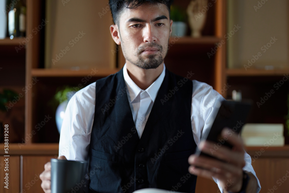 A portrait of a handsome and discreet Asian man sitting at his desk using his phone while taking a break from reading and drinking coffee A portrait of a handsome and discreet Asian man sitting at his desk using his phone while taking a break from reading and drinking coffee