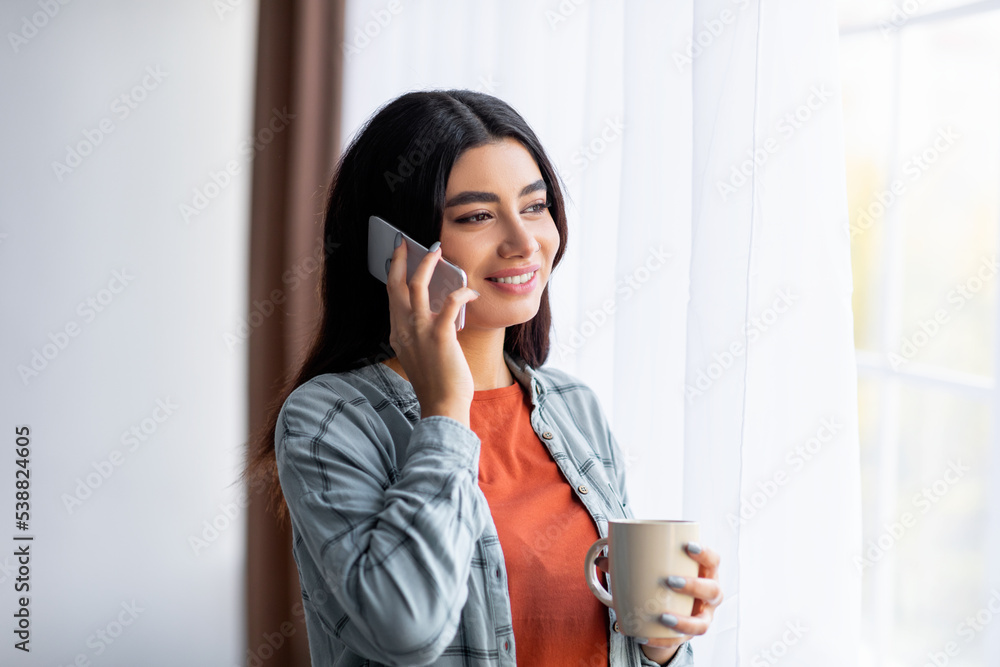 Happy young Arab woman standing near window with coffee, speaking on smartphone, communicating from home Happy young Arab woman standing near window with coffee, speaking on smartphone, communicating from home