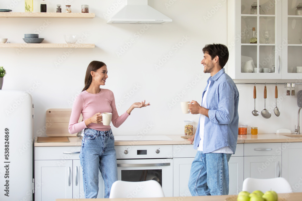 Fototapeta premium Smiling relaxed millennial caucasian woman and man with cups of drink talk on kitchen interior