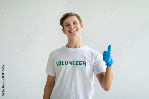 Volunteer work. Happy activist guy in white volunteer t-shirt showing thumb up, posing and smiling over light background