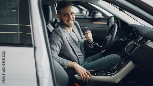 Close-up portrait of handsome bearded guy new car owner sitting inside beautiful automobile holding key fob and smiling looking at camera. Transportation and people concept.