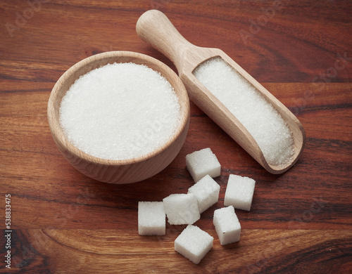 Miseczka i łopatka z białym cukrem, obok duże kostki białego cukru na drewnianym tle