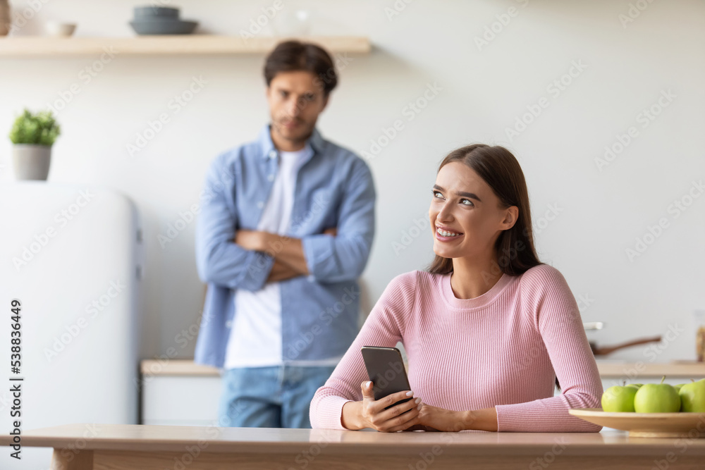 Offended sad millennial male looks at smiling female with smartphone at kitchen Offended sad millennial male looks at smiling female with smartphone at kitchen