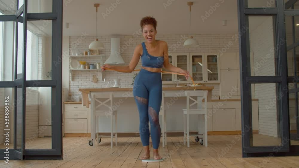 Happy smiling charming black woman in activewear with measuring tape standing on weight scale, cheering successful slimming and making dancing moves, showing excitement and joy indoors. Happy smiling charming black woman in activewear with measuring tape standing on weight scale, cheering successful slimming and making dancing moves, showing excitement and joy indoors.