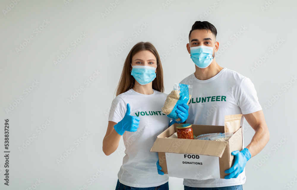 Volunteer work. Young happy volunteers in medical masks holding food donations box, woman showing thumb up, copy space Volunteer work. Young happy volunteers in medical masks holding food donations box, woman showing thumb up, copy space
