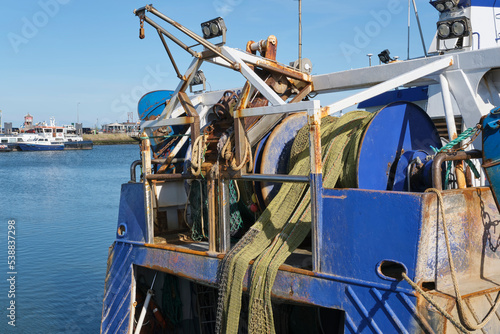 Rolled up industrial fishing net on a winch aboard a rusty blue fishing boat or trawler under a blue sky. 