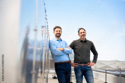 Smiling mature businessman with arms crossed standing by colleague on rooftop photo