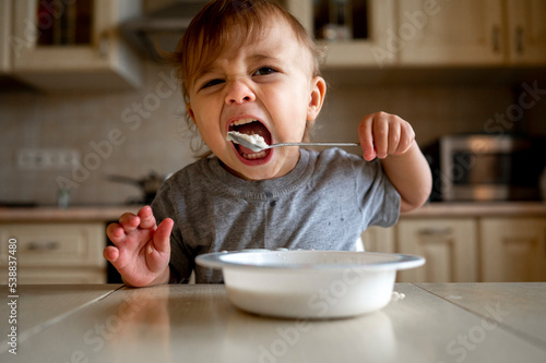 Baby boy eating porridge with spoon in kitchen