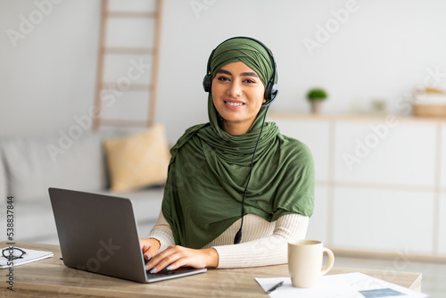 Canvastavla Young Arab female in hijab working on laptop from home, wearing headphones, usin