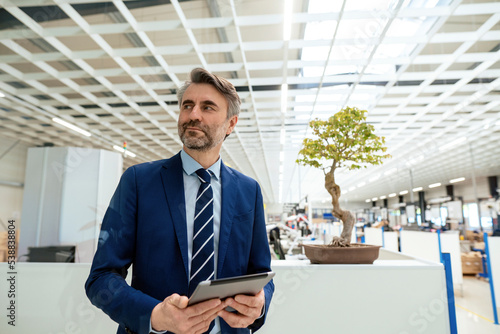 Smiling mature businessman with tablet PC standing in front of bonsai tree at industry photo