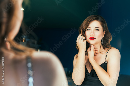 Portrait of posh woman with makeup and stylish dark hair in elegant cocktail silk black dress looking on herself in mirror reflection. Fashionable and self-confident lady. Self Assurance