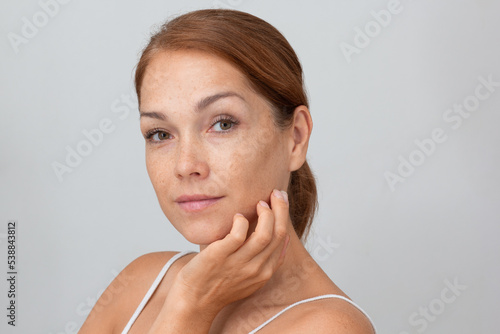 Portrait of cropped caucasian middle aged woman face with freckles holding fingers on cheek, showing soft skin on white background looking at camera photo