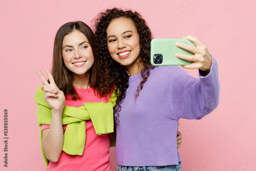Young two friends women wear green purple shirts looking camera together doing selfie shot on mobile cell phone post photo on social network show v-sign isolated on pastel plain light pink background. Young two friends women wear green purple shirts looking camera together doing selfie shot on mobile cell phone post photo on social network show v-sign isolated on pastel plain light pink background.