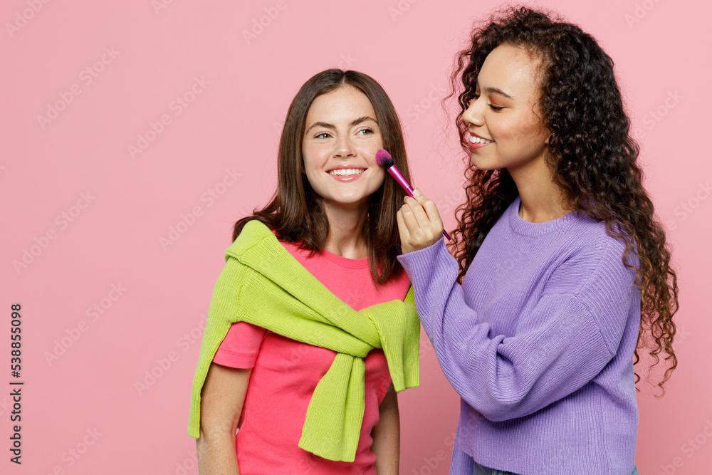Young two women 20s wear green purple shirts together do make up for friend at beauty salon use brush for applying blush or powder isolated on pastel plain light pink color background studio portrait. Young two women 20s wear green purple shirts together do make up for friend at beauty salon use brush for applying blush or powder isolated on pastel plain light pink color background studio portrait.