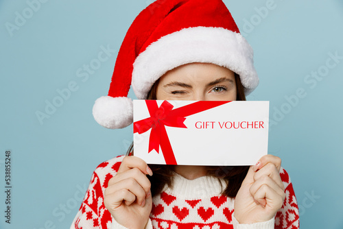 Young merry woman wear red sweater Santa hat posing cover mouth gift certificate coupon voucher card for store isolated on plain pastel light blue cyan background. Happy New Year 2023 holiday concept.