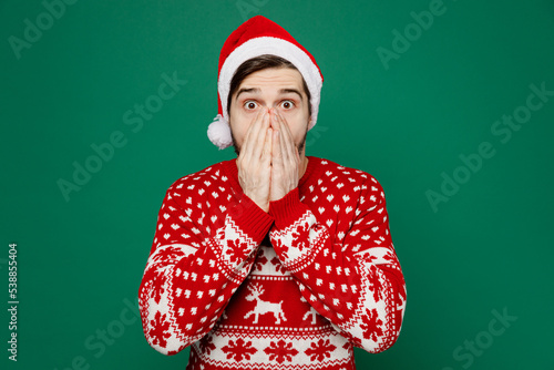 Merry young man 20s wear red warm knitted sweater Santa hat posing cover mouth with hand look camera isolated on plain dark green background studio portrait. New Year 2023 holiday celebration concept.
