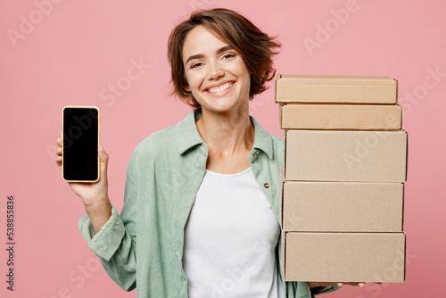 Young woman wear green shirt white t-shirt hold in hand use mobile cell phone with black screen area brown clear blank craft paper cardboard box mock up isolated on plain pastel light pink background