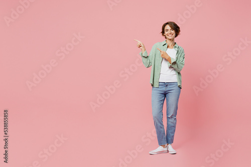 Full body young happy woman 20s she wear green shirt white t-shirt point index finger aside on workspace area isolated on plain pastel light pink background studio portrait. People lifestyle concept.