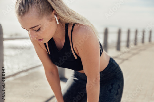 Blonde female runner resting bent over on promenade after run along the beachfront on a summer afternoon
