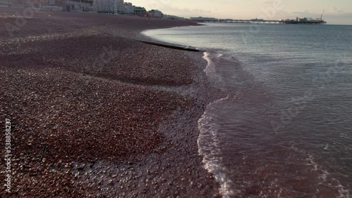 Drone flies back and low over Brighton Beach at dawn in slow motion. Palace Pier in background