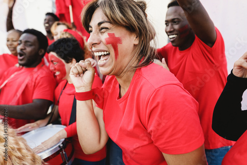 Multiracial red sport fans screaming while supporting their team - Football supporters having fun at competition event - Soft focus on latin woman face