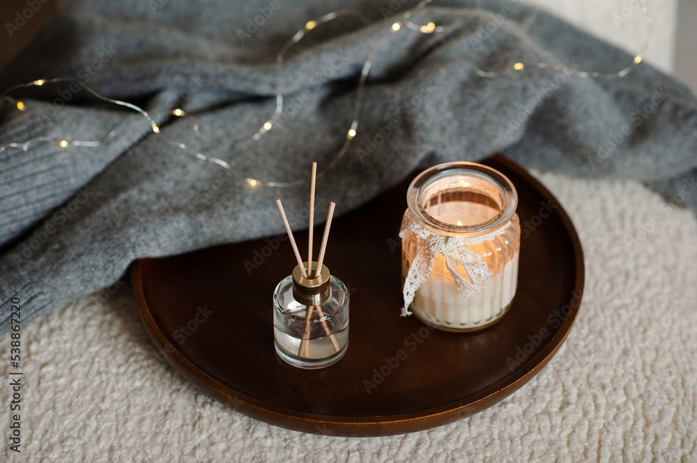 Home liquid perfume in glass bottle and bamboo sticks with scented candle with knitted sweater on wooden tray in cozy chair. Aromatherapy. Home liquid perfume in glass bottle and bamboo sticks with scented candle with knitted sweater on wooden tray in cozy chair. Aromatherapy.