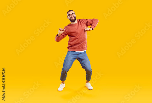 Happy male dancer or fashion model in casual outfit dancing on yellow studio background. Funny cheerful young man in orange sweatshirt, blue jeans and glasses dancing and having fun
