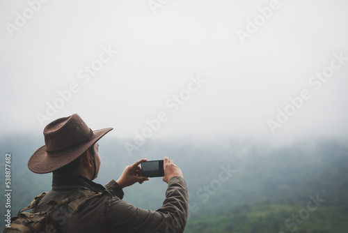 Freedom traveler man in hat carrying a backpack stands at the top of a mountain and taking photo by smartphone on a foggy day.Adventure travel and success concept