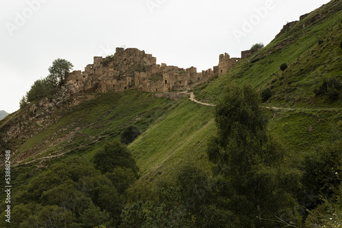 Ancient ruin of stone impregnable, destruction city on peak of mountain in highlands in Dagestan with green slope, old dirt road in summer, in overcast. Breathtaking adventure and travel in mountain.