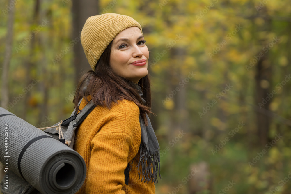 Woman hiker with smiling and excited expression enjoys hiking and exploring nature of autumn forest. Lady tourist with backpack turns around looking at trees, copyspace