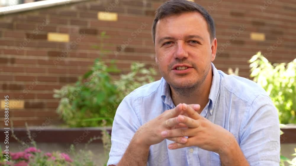 Millennial confident Caucasian man explains to camera as he sits on street in urban area with apartment building in background. Man blogger talking to camera with his audience outdoors.