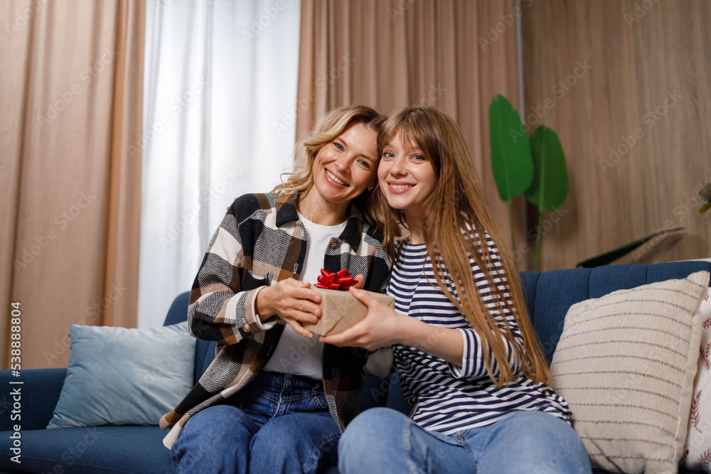 Portrait of happy mother and her daughter with gift box on sofa in living room. Young woman gives a present to her mom Portrait of happy mother and her daughter with gift box on sofa in living room. Young woman gives a present to her mom