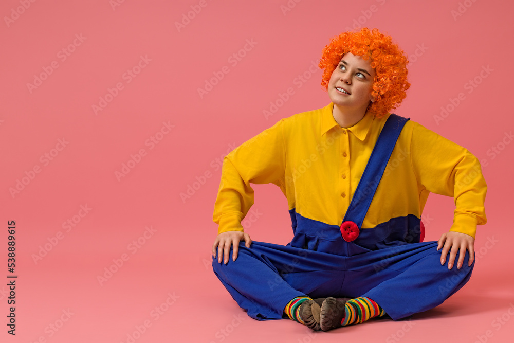 cheerful clown in a wig and a yellow-blue suit sits cross-legged and looks up on a colored background with copy space cheerful clown in a wig and a yellow-blue suit sits cross-legged and looks up on a colored background with copy space