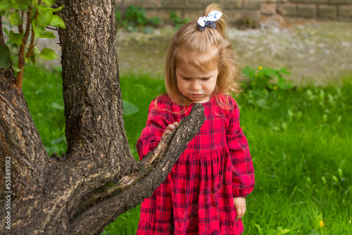 A little sad girl in a red checkered dress near a tree