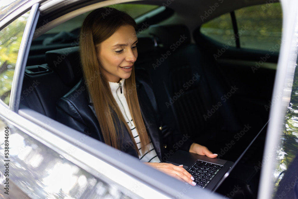 Beautiful business woman is using a laptop and smiling while sitting on back seat in the car Beautiful business woman is using a laptop and smiling while sitting on back seat in the car