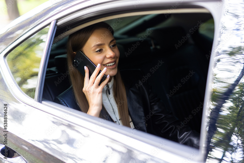 Beautiful woman is talking on the mobile phone, using a digital tablet and smiling while sitting on back seat in the car Beautiful woman is talking on the mobile phone, using a digital tablet and smiling while sitting on back seat in the car